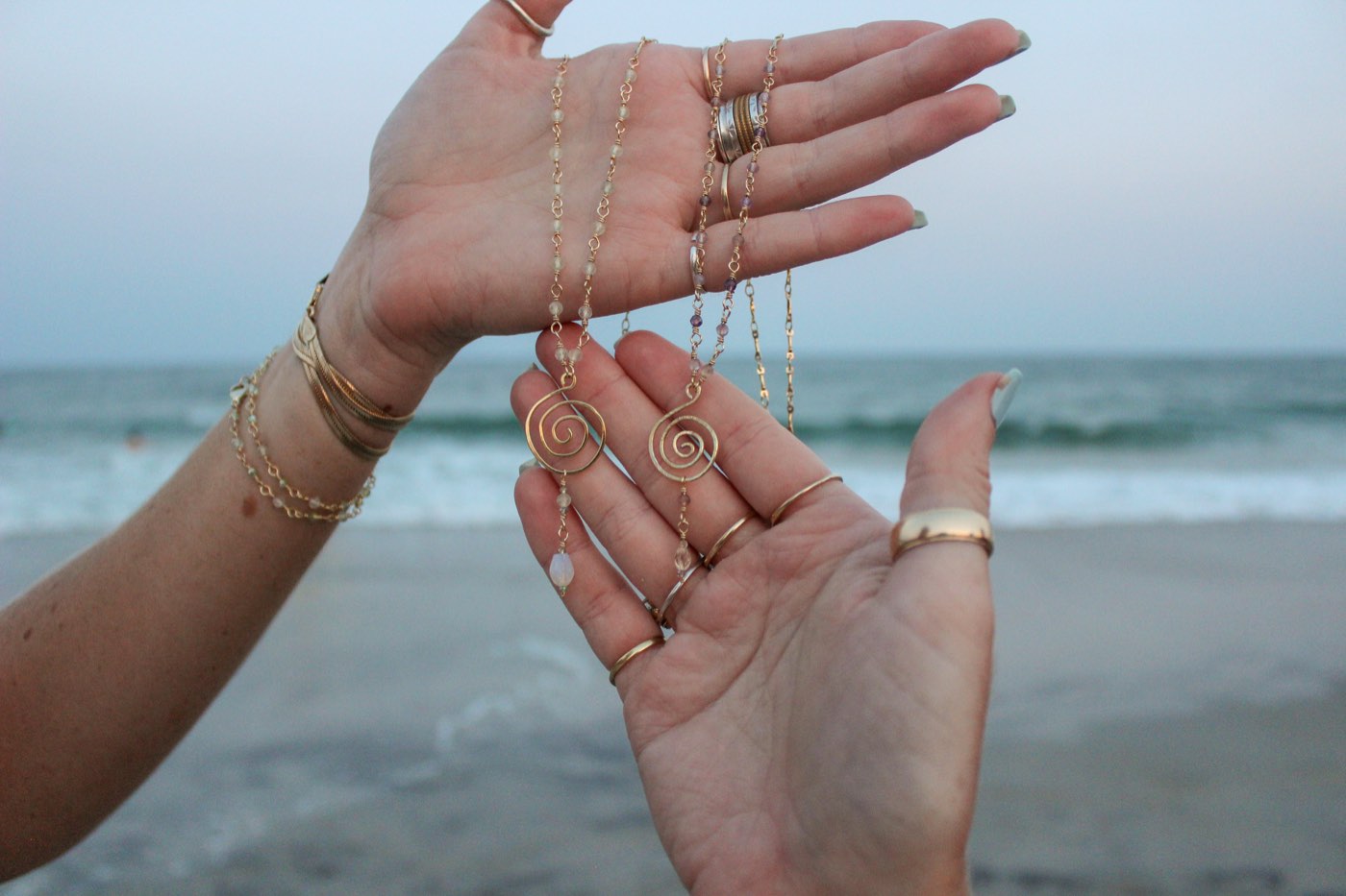 Two hands holding spiral pendants on beaded chains at the shore